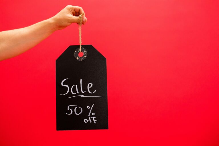A red background with a hand holding a black sale tag reading 50% off — symbolic image for consumer desensitization to discounts.