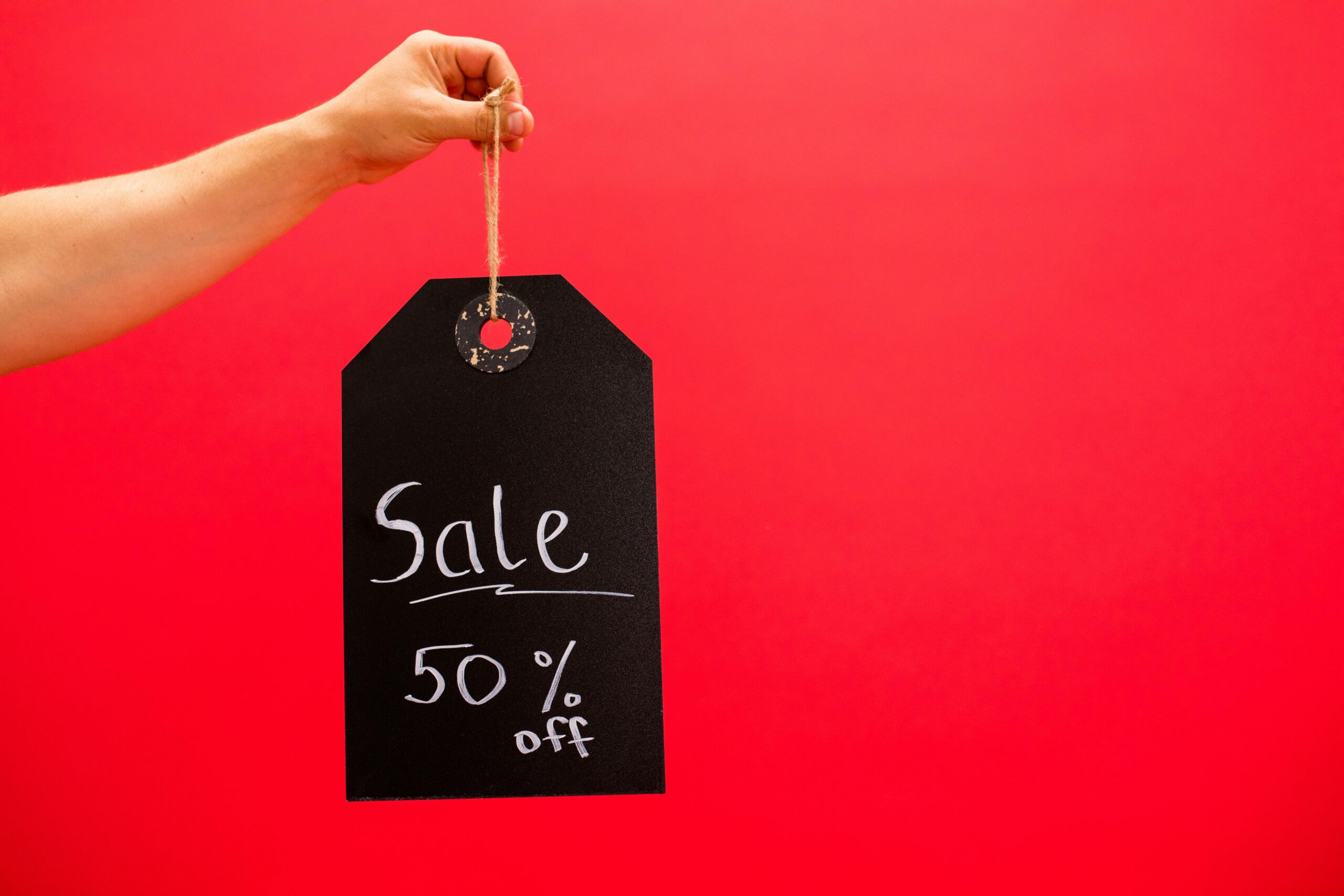 A red background with a hand holding a black sale tag reading 50% off — symbolic image for consumer desensitization to discounts.